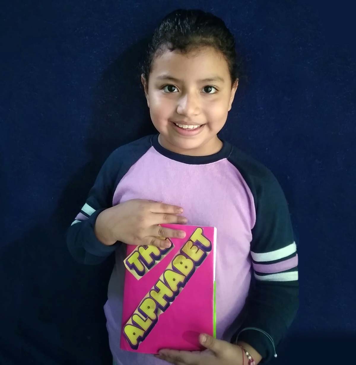 Kamila wearing purple long-sleeve sweater with mint-green stripes on the arms and holding pink book with the title, The Alphabet