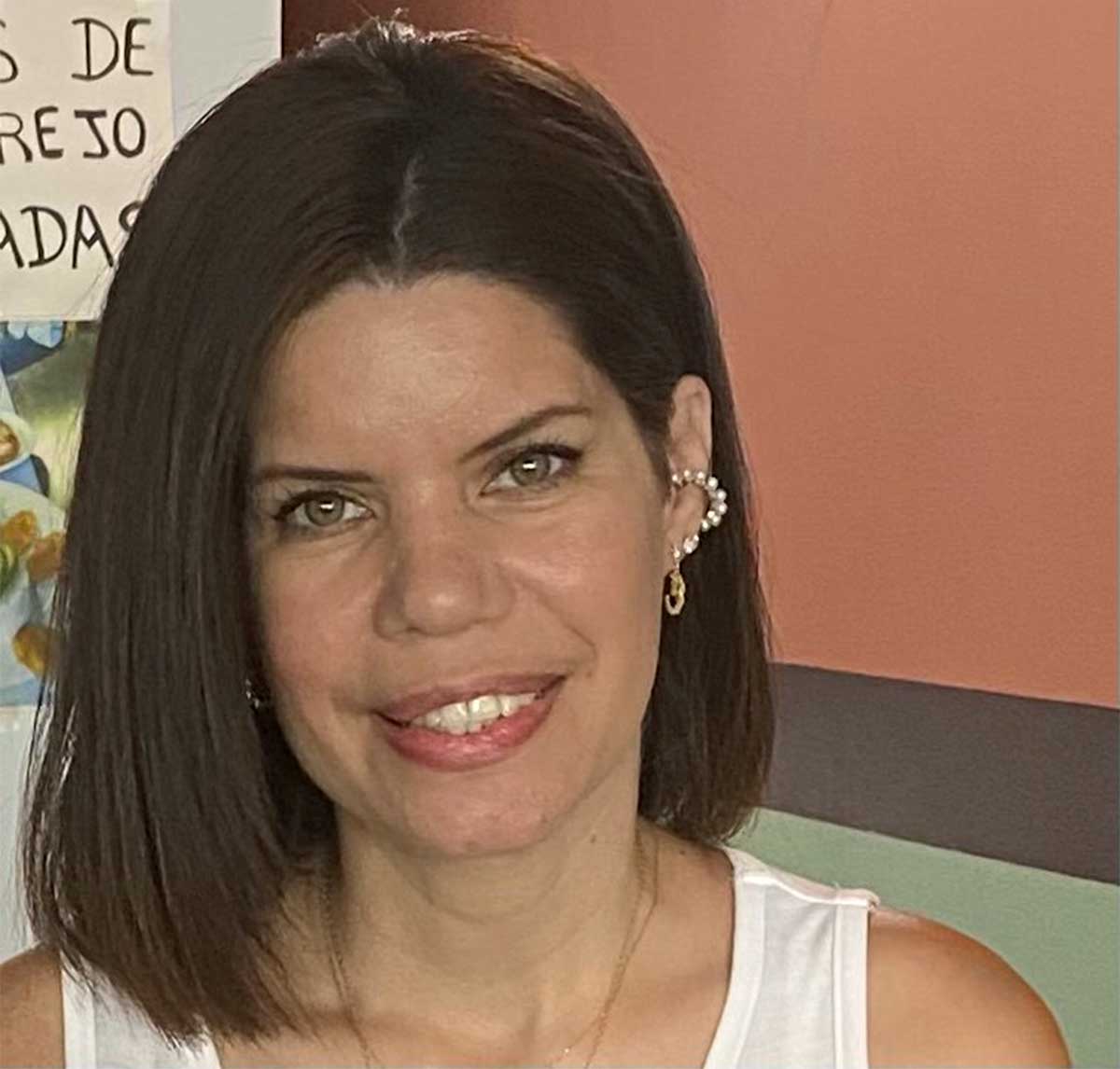 Madelyn Cardenas, a smiling woman with shoulder-length brown hair, green eyes, pearl ear cuff earrings, white sleeveless top.