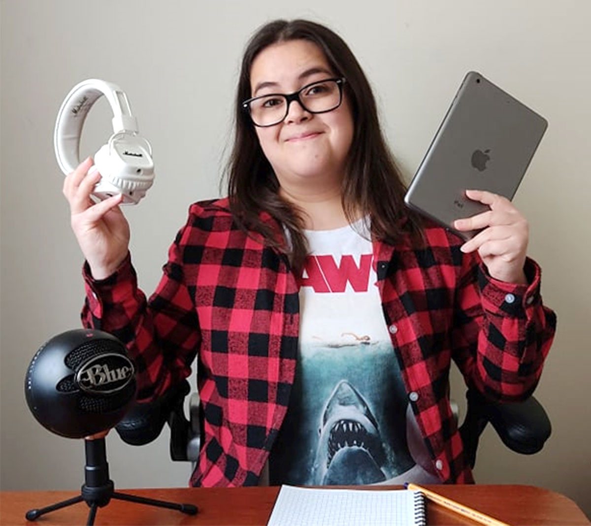 Camila Muñoz, a smiling woman with long hair, glasses, and a plaid shirt sitting at her desk holding an Ipad and headphones.